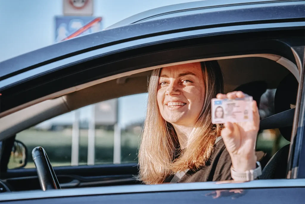 Glückliche Frau zeigt stolz ihren neuen Führerschein aus dem Auto – Erfolgreiche Führerscheinausbildung bei der Fahrschule Müller.